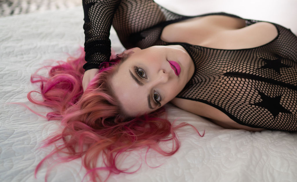 woman with bright pink hair laying on a bed in a black bodysuit NKD Worcester